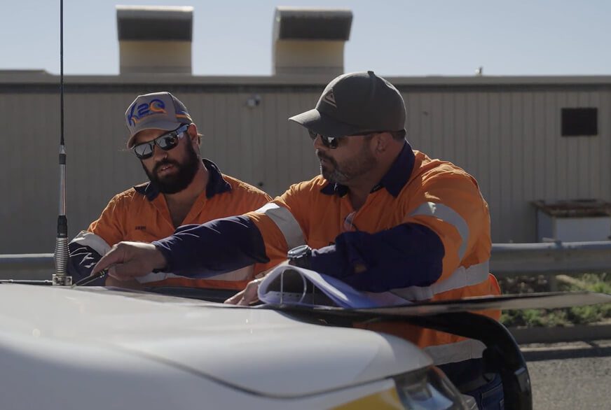 Two workers in orange hi-vis reviewing plans on the bonnet of a site vehicle.