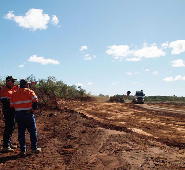 Two workers in orange hi-vis standing on a large earthworks site while machinery and a truck operate in the background.