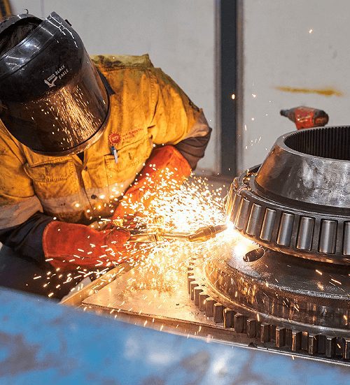 Tradesperson in welding helmet working on a large gear component with bright sparks.